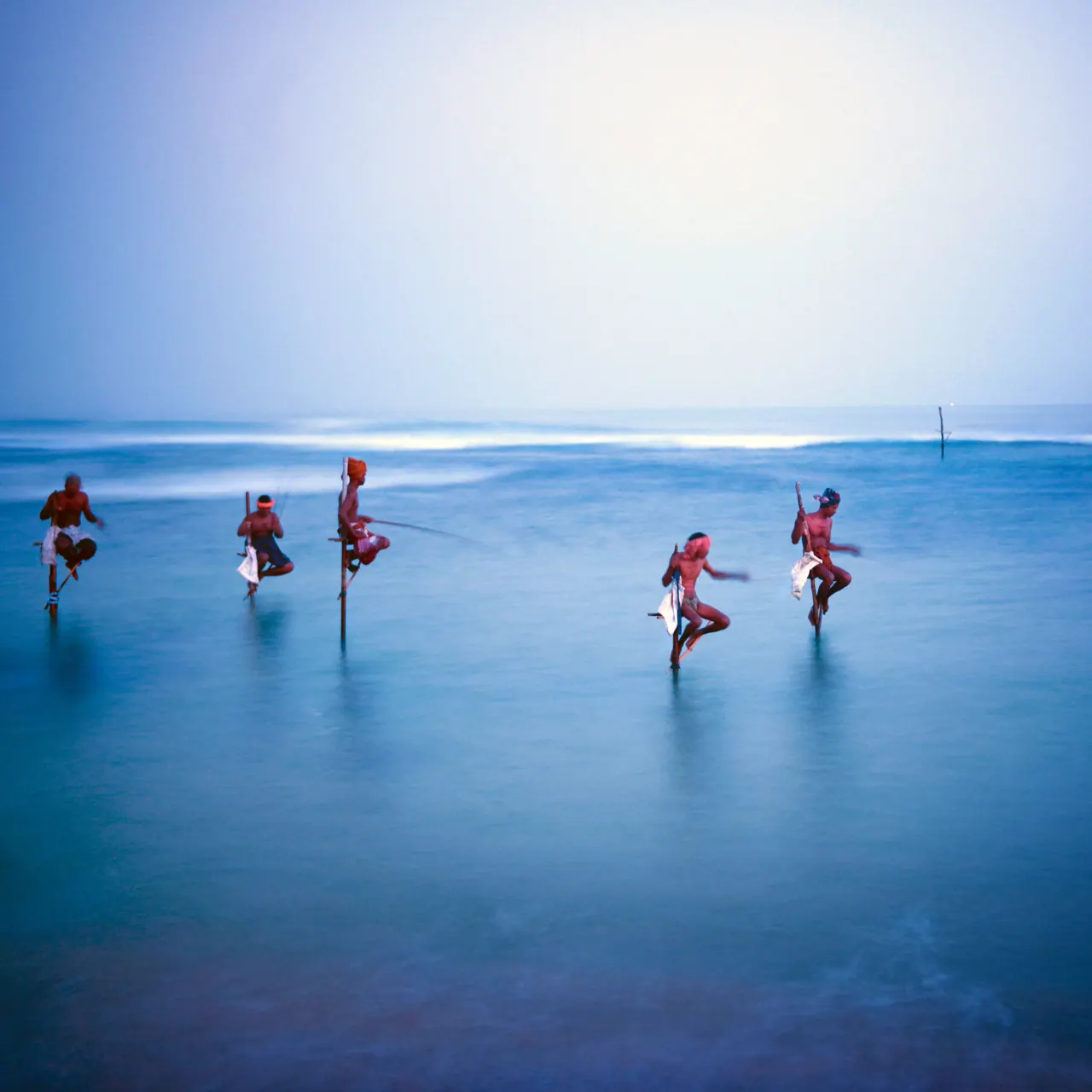 Stilt fishermen perched on poles above a calm sea at dusk